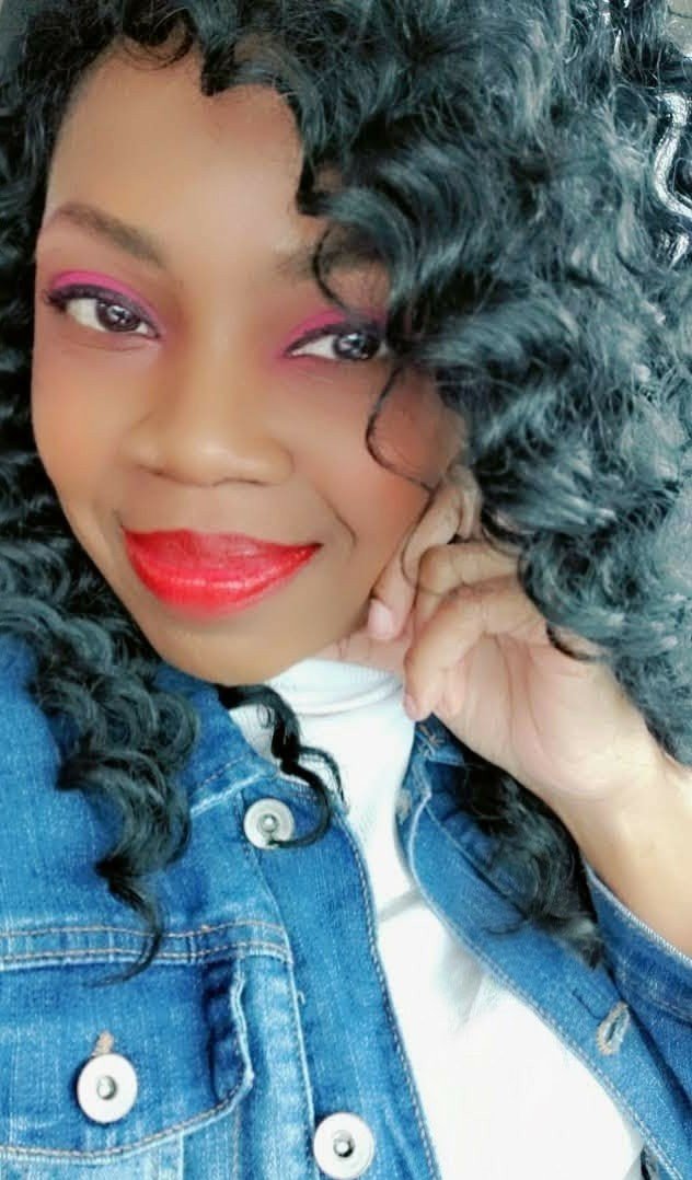 Nisha Vee smiling in a denim jacket with bold pink eyeshadow and red lipstick